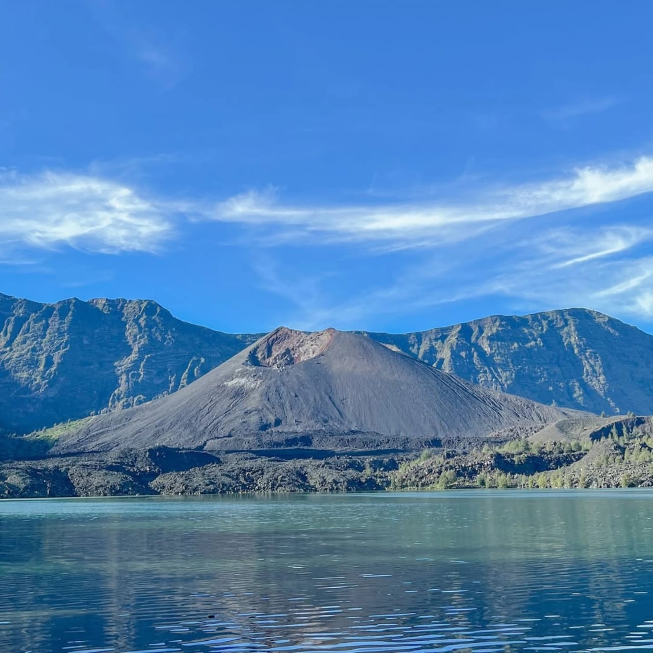 A scenic view of the Segara Anak caldera lake inside Mount Rinjani, Lombok, Indonesia. The clear blue-green water reflects the surrounding mountains. The active, cone-shaped Mount Barujari volcano sits in the center, with steep caldera walls in the background under a bright blue sky with wispy clouds.